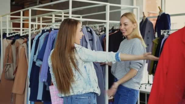 Hermosa mujer joven está eligiendo abrigo en la tienda y conocer a una amiga. Las chicas están felices de verse, se están abrazando, riendo y charlando . — Vídeos de Stock