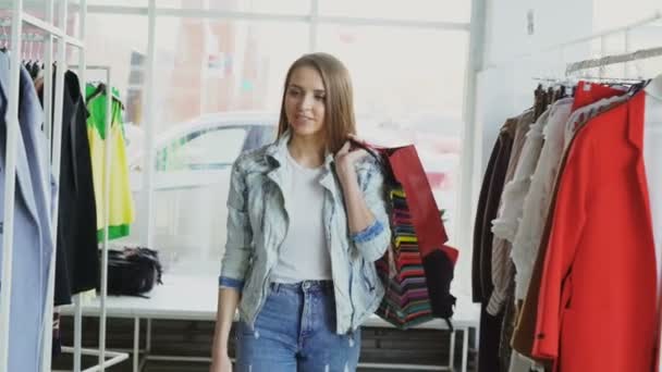 Dolly shot of attractive young lady walking slowly through modern shop. Elle tient plusieurs sacs en papier et regarde de beaux vêtements et chaussures exposés autour . — Video