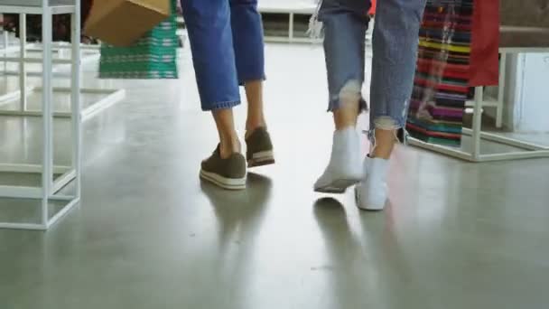 Vista posterior de las piernas de las mujeres que van lentamente a través de una amplia tienda. Las niñas llevan pantalones vaqueros y zapatillas y bolsas de papel de colores. . — Vídeos de Stock