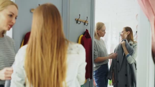 Dość młoda kobieta jest sprawdzanie modny płaszcz w Przymierzalnia z przyjaciółką, pomagając jej oceniać odzieży. Są rozmowy, gestykulacji i patrząc na ubrania. — Wideo stockowe