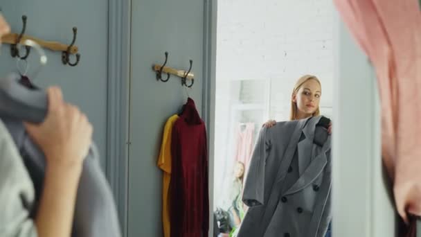 Ελκυστική νεαρή γυναίκα προσπαθεί κομψό παλτό ενώ στέκεται σε δοκιμαστήρια είναι boutique ρούχα. Αυτή κοιτάζοντας τον εαυτό της στον καθρέφτη, στροφή και χαμογελαστός. — Αρχείο Βίντεο
