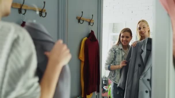 Giovane donna allegra sta cercando cappotto in camerino e chiedendo al suo amico per un consiglio. Fidanzata si avvicina e si stanno guardando allo specchio insieme e sorridendo felicemente . — Video Stock