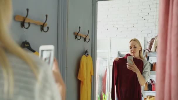 Cliente feminino está fazendo selfie espelho com novo jumper de malha usando telefone inteligente. Ela está de pé no vestiário na loja de roupas e posando com um lindo sorriso . — Vídeo de Stock