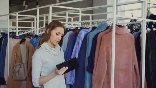 Молода бізнес-леді перевіряє одяг у своєму магазині з планшетом в руках. Її працівник приходить і питає про одяг. Роботодавець розмовляє з нею і жестикулює . — стокове відео