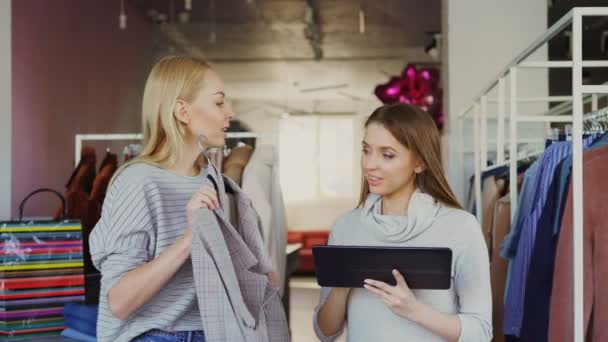 Jovem empresário está usando tablet enquanto está de pé em sua loja de roupas. A assistente dela vem com roupa e pede conselhos. Mulher de negócios está mostrando-lhe onde colocá-lo . — Vídeo de Stock