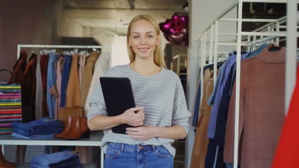 Retrato de sucesso jovem empresária em pé em sua loja de roupas, segurando tablet, sorrindo e olhando para a câmera. Boutique espaçoso com roupas femininas no fundo . — Vídeo de Stock