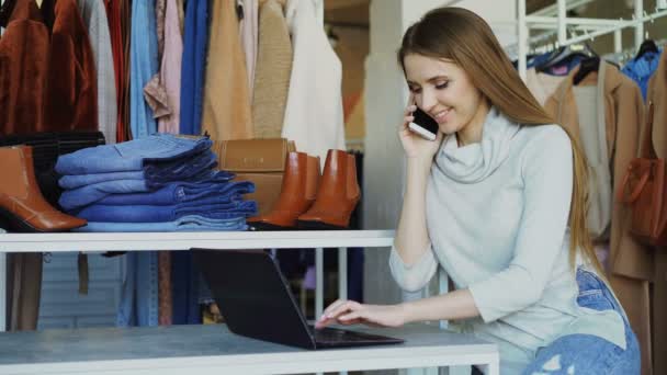 Giovane donna d'affari sta lavorando con il computer portatile e parlando sul cellulare nel suo negozio. Abbigliamento e cliente in background. Concetto di piccola impresa . — Video Stock