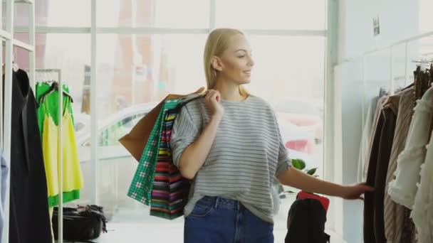 Dolly tiro de menina loira andando entre prateleiras e trilhos em grande loja. Ela está carregando lotes de sacos, sorrindo e olhando para roupas finas em torno dela . — Vídeo de Stock