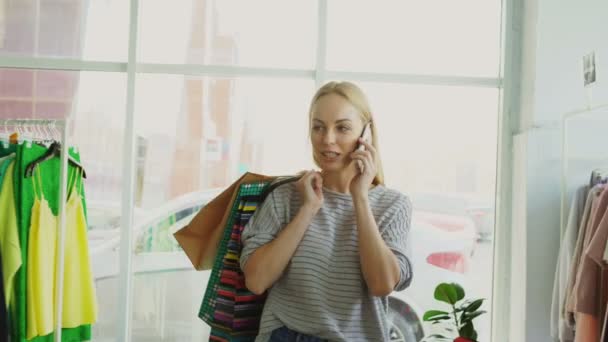 Attractive blond woman is walking between shelves and rails in large store and talking on mobile phone. She is carrying bags, smiling and looking at trendy clothes around her. — Stock Video