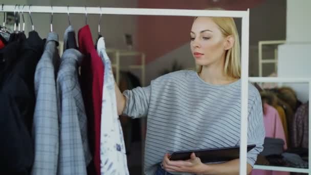 Владелец магазина одежды проверяет и подсчитывает одежду на рельсах при работе с планшетом. Она вводит информацию о своих товарах. Концепция малого бизнеса . — стоковое видео