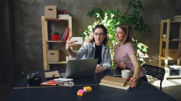 Jóvenes empresarias están haciendo selfie juntos en la oficina moderna mientras están sentadas en el escritorio. Ellos están posando con caras divertidas y sonriendo, luego viendo fotos . — Vídeos de Stock