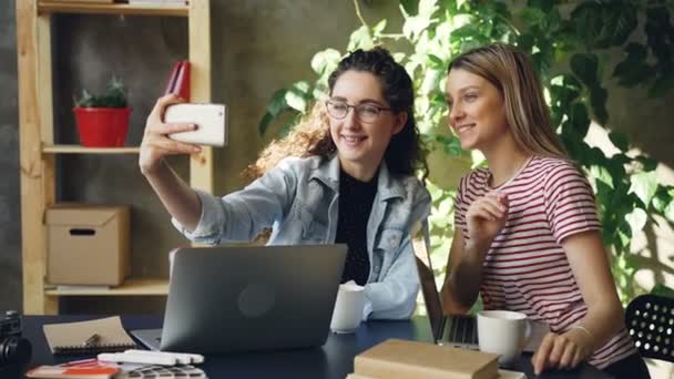 Los jóvenes emprendedores están hablando y haciendo selfie con teléfono inteligente mientras están sentados en el escritorio en la oficina moderna. Están posando y riendo, luego viendo fotos . — Vídeos de Stock