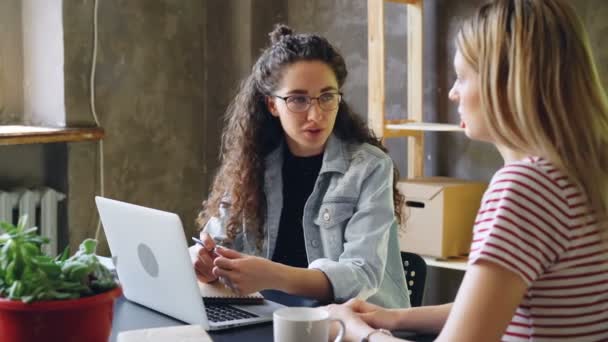 La joven emprendedora está hablando con su pareja en la oficina moderna mientras está sentada en el escritorio juntos. Las mujeres están planeando un nuevo proyecto. Concepto de cooperación exitosa . — Vídeo de stock