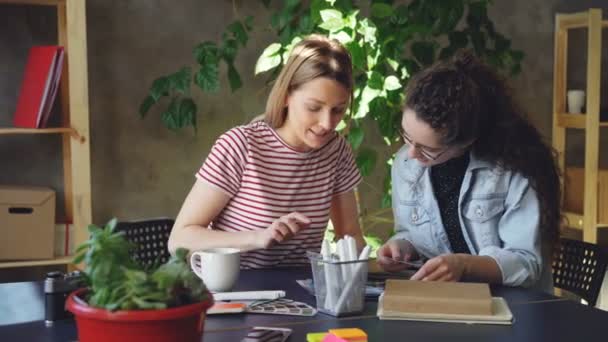 Equipe de dois designers criativos estão olhando para fotos e falando sobre eles enquanto se sentam juntos na mesa. Panela de tiro de escritório informal moderno. Ambiente de trabalho amigável . — Vídeo de Stock