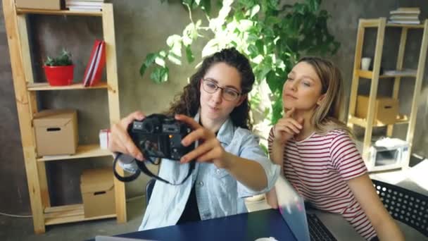 Las estudiantes femeninas están haciendo selfie con la cámara mientras se sientan juntas en la mesa en la oficina moderna. Ellos están posando, sonriendo y tirando caras divertidas . — Vídeo de stock