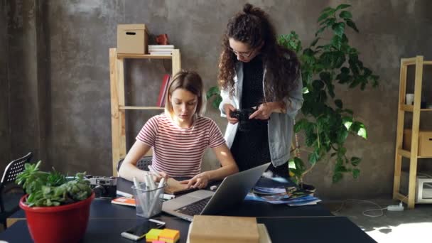 Desainer muda menggambar gambar di notebook duduk di meja sementara fotografer wanita datang kepadanya dengan kamera. Perempuan mulai membahas proyek dan menonton foto di layar . — Stok Video