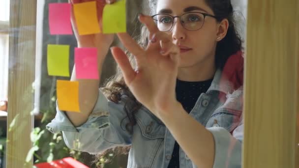 Close-up of young attractive woman wearing glasses sticking colored memos on glass board in modern office. She is looking at bright notes and smiling. — Stock Video