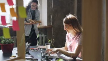 İki çekici kızlar birlikte modern loft ofiste çalışıyoruz. Sarışın kadın onun meslektaşım kitap ayakta pencere yakınındaki okurken masada oturan dizüstü bilgisayarda yazarak
