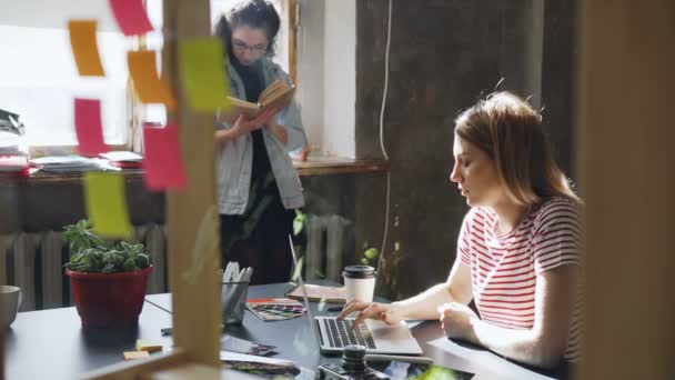 Fiatal szőke diák alkalmi ruhában elfoglalva, Laptop gépelés, miközben barátja van olvasó könyv állandó ablak közelében. Loft stílusú lakás van a háttérben. — Stock videók