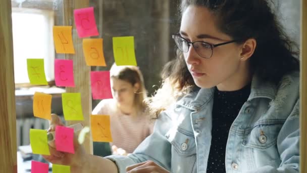 Jeune femme d'affaires créative numérotant des autocollants colorés sur verre pour le projet tout en travaillant avec une collègue dans un bureau moderne — Video