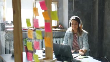 Yaratıcı genç işkadını modern ofis içinde laptop ile masasında çalışırken dans kulaklık müzik dinliyor. Cam ön planda renkli çıkartmaları ile.