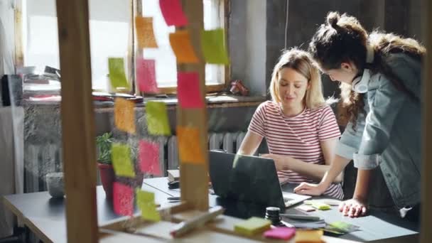 El equipo creativo de diseñadoras está trabajando en un nuevo proyecto, compartiendo ideas, escribiendo información sobre pegatinas de colores. La pared de la oficina de cristal con memos brillantes está en primer plano . — Vídeo de stock