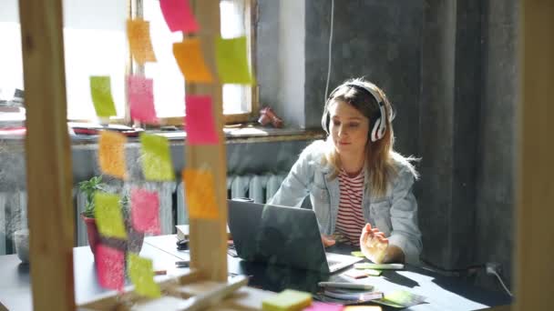 A csinos szőke lány zenét hallgatni, és a laptop szép loft hivatalban dolgozó. Ő így megjegyzi a Jegyzettömb, mosolyogva, és a tánc. Kellemes munkahelyi koncepció. — Stock videók