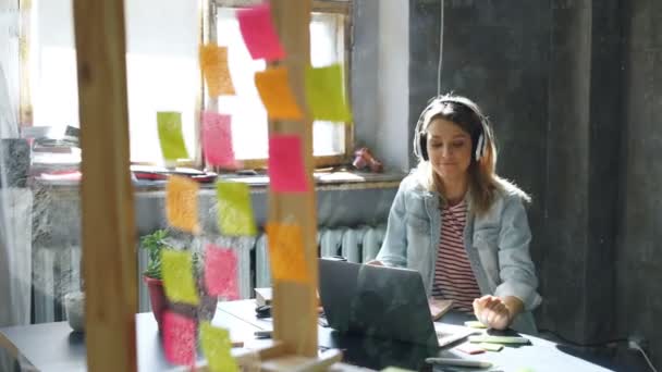 Kreatív fiatal üzletasszony hallgatni zene, tánc, miközben dolgozik a laptop a modern irodai íróasztal fejhallgató. Üveg előtérben színes matricával. — Stock videók