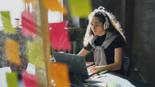 Aantrekkelijke vrouwelijke student is muziek beluisteren met koptelefoon dansen en zingen terwijl het werken met een laptopcomputer. Glassboard met helder gekleurde stickers op voorgrond. — Stockvideo