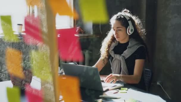 Creatieve jonge zakenvrouw is luisteren naar muziek in hoofdtelefoons dansen en zingen terwijl hij werkte bij Bureau met laptop in moderne kantoor. Glas met gekleurde stickers op voorgrond. — Stockvideo