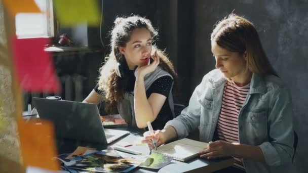 Cheerful girl is working with laptop and listening to music, then taking off headphones and talking with her coworker and laughing. Productive teamwork concept. — Stock Video