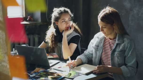 Chica alegre está trabajando con el ordenador portátil y escuchar música, a continuación, quitarse los auriculares y hablar con su compañero de trabajo y riendo. Concepto de trabajo en equipo productivo . — Vídeos de Stock