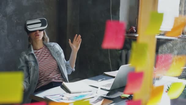 Mujer joven atractiva está sentada en la mesa y con gafas de realidad virtual. Ella está moviendo las manos y y teniendo experiencia VR en lof oficina moderna — Vídeos de Stock