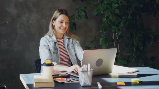 有魅力的年轻女子坐在现代办公室的桌子上, 在笔记本电脑上谈论 skype。她在说感情, 示意和笑. — 图库视频影像