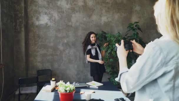 Приваблива дівчина позує з великою рослиною, а колега фотографує її на цифровій камері в сучасному офісі. Жінки розважаються і сміються під час перерви на каву — стокове відео