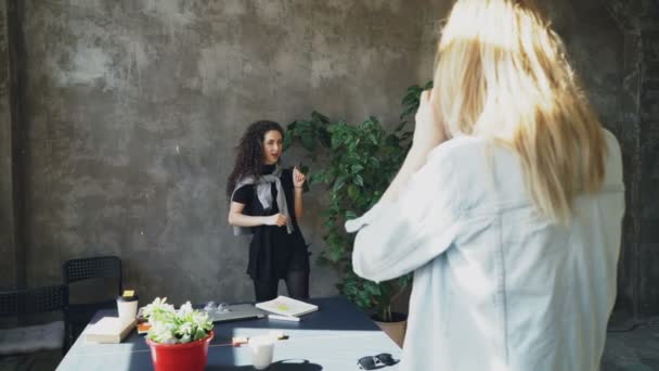Attraktives Mädchen posiert mit großer Pflanze, während eine Kollegin sie mit der Digitalkamera im modernen Büro fotografiert. Frauen haben Spaß und lachen in der Kaffeepause — Stockvideo