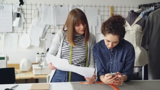 Mladí oblečení design podnikatelé jsou při pohledu na skici a kontrola smartphone. Atraktivní ženy jednají módního průmyslu v přátelsky. — Stock video