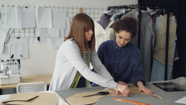 La costurera experimentada está enseñando a su asistente a delinear patrones de ropa en la tela. Mujer joven se centra en el proceso, observando cuidadosamente y haciendo preguntas . — Vídeos de Stock