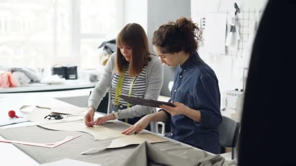 Attractive clothes designer entrepreneurs are outlining cutout with chalk, working with tablet and talking. Busy day at successful tailors shop concept. — Stock Video