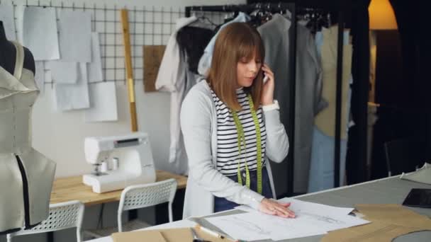 Die attraktive Schneiderin bestellt per Handy Stoff für ihre neue Kleiderkollektion und betrachtet Skizzen mit kleinen Details. Kommunikation mit Lieferantenkonzept. — Stockvideo