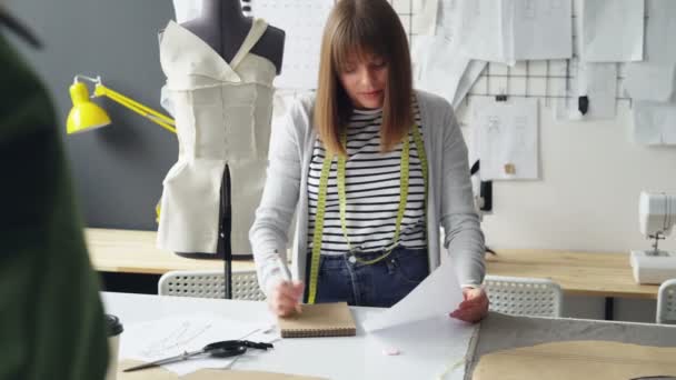 Dressmaker profesional está tomando notas en bloc de notas y mirando el bosquejo de ropa mientras se trabaja en la mesa del estudio. Muchos dibujos, maniquí y máquina de coser en el fondo . — Vídeos de Stock