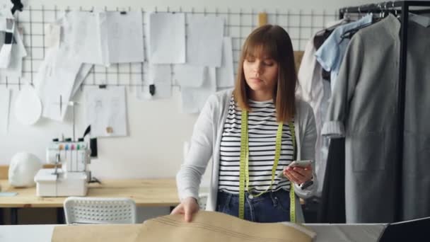 Abbigliamento imprenditore di design sta controllando bozze di carta da cucire e guardando smartphone. Sta esaminando attentamente ogni ritaglio e pensando a un indumento futuro. . — Video Stock