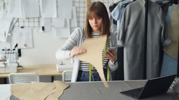 Ung sömmerska kontroll kläder papper mönster och mäta dem med måttband medan du tittar på smart telefon. Trevlig studio med plagg och symaskin i bakgrunden. — Stockvideo