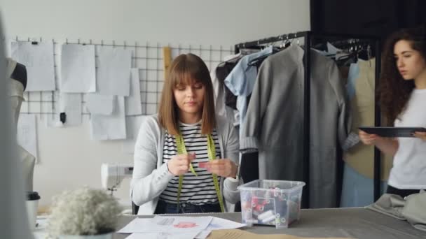 La empleada de la sastrería está midiendo dibujos de prendas con hilo de coser mientras su colega muestra la pantalla de su tableta y habla con ella. . — Vídeos de Stock