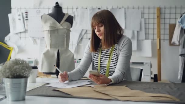 Diseñador de ropa creativa está buscando en el teléfono inteligente y dibujo bosquejo mientras trabaja en la sastrería moderna en la mesa de coser. La mujer está ocupada e involucrada en el proceso . — Vídeos de Stock