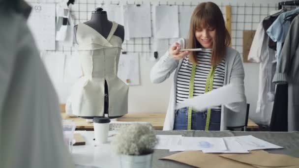 Ambicioso sastre creativo femenino está colocando bocetos de prendas de vestir en el escritorio del estudio y disparándoles con un teléfono inteligente. Presentación de dibujos de nuevo concepto de colección . — Vídeo de stock