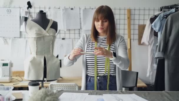 Beautiful woman fashion designer is taking photos of sketches lying on table. She is making creative flat lays. Her studio is light and full of sewing items, tools and equipment. — Stock Video