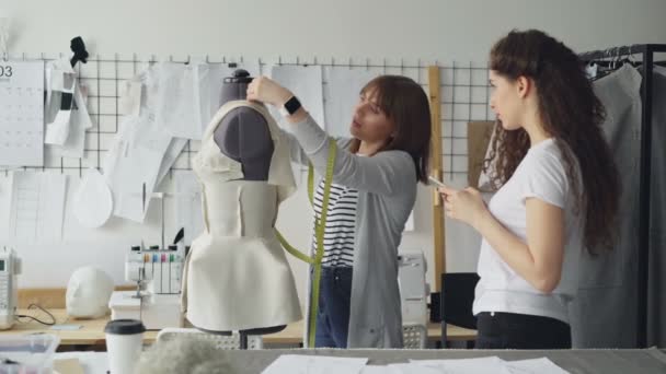 Jeune femme mesure des pièces de mannequin avec ruban à mesurer tandis que son collègue saisit cette information dans son smartphone. Coopération productive dans l'adaptation du concept d'entreprise . — Video