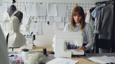 Profesyonel kadın terzi dikiş makinesi ayarlama sonra kumaş parçaları dikiş. Kadın studio terzilik ışıkta çalışıyor. Dikiş öğeleri ve mankenler tarafından görülebilir.