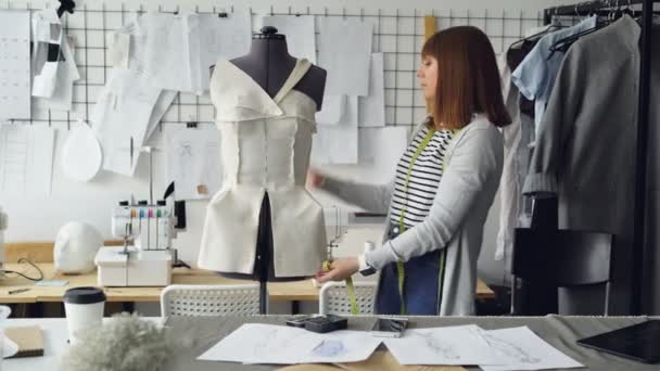 Self-employed female tailor is checking clothing patterns pinned to tailoring dummy then taking photos of it with smart phone. Nice workshop with sewing machines in background. — Stock Video